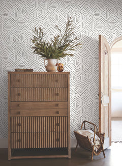 A wooden dresser with striped drawers stands next to an open arched door. A vase with greenery sits on top, and a woven basket with blankets is on the floor. The wall features the Sirih Wallpaper by York Wallcoverings, showcasing a geometric black-and-white pattern.