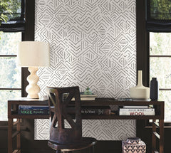 A wooden desk adorned with books, a lamp, and ceramics is positioned in front of a window dressed with York Wallcoverings’ Sirih Wallpaper. A dark wooden chair faces the desk as natural light filters through the window.