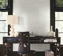 A wooden desk adorned with books, a sculpted lamp, and vases sits against York Wallcoverings’ Sirih Wallpaper between two windows, with a carved wooden chair placed in front.