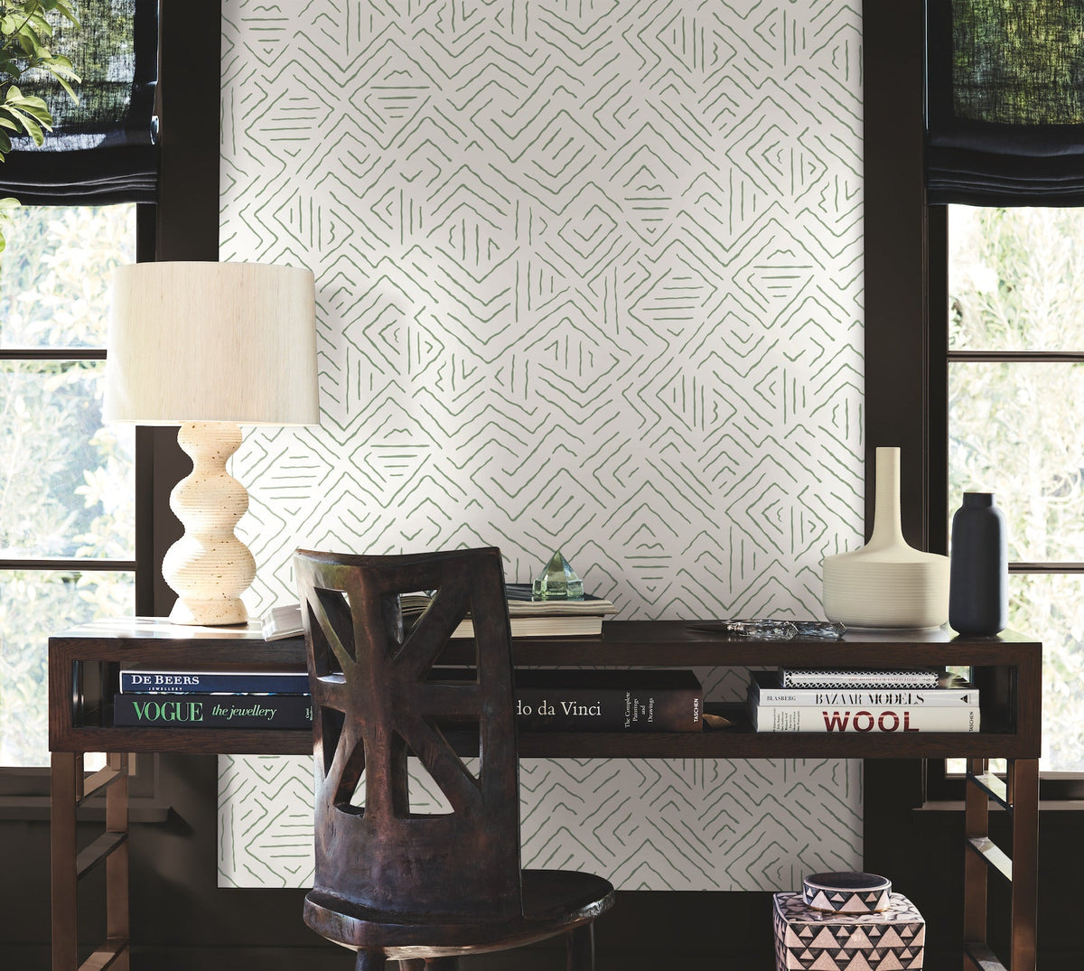A wooden desk with books, a lamp, and decorative vases sits in front of two windows. A dark chair is pushed in, and the wall behind the desk is adorned with York Wallcoverings’ Sirih Wallpaper.