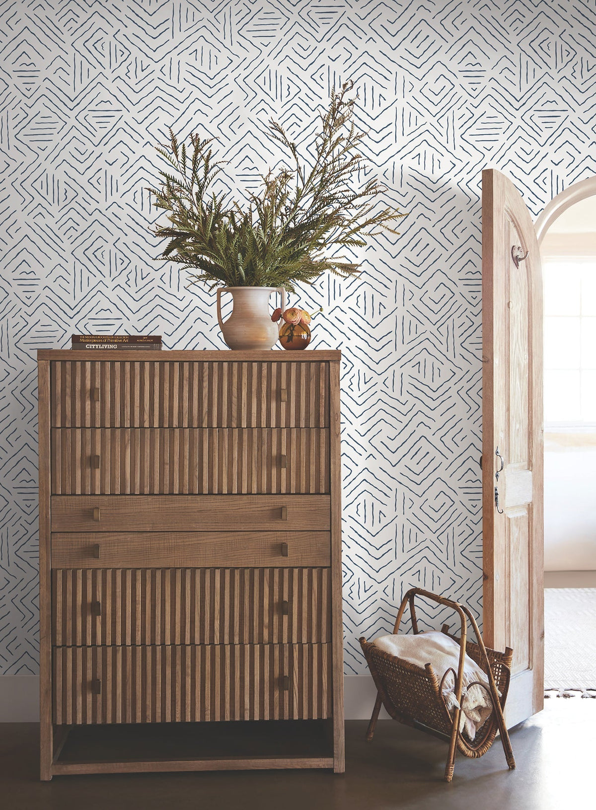 A wooden chest of drawers with a striped pattern stands against York Wallcoverings’ Sirih Wallpaper. A vase with greenery sits on top, while a basket filled with blankets rests on the floor near an arched doorway letting in light.