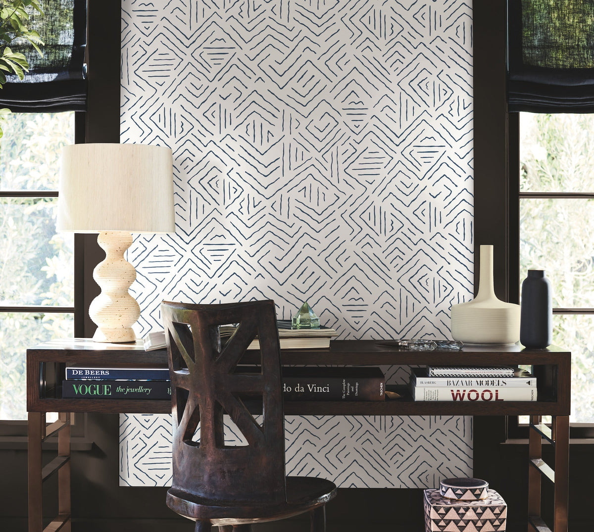 A wooden desk topped with books, a lamp, and ceramic vases sits against a wall adorned with York Wallcoverings’ Sirih Wallpaper. The geometric pattern is framed by two windows featuring black shades. A carved wooden chair is positioned in front of the desk.