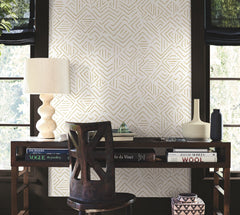 A wooden desk with books, a lamp, and vases sits by two windows, paired with a dark chair facing it. The wall is adorned with Sirih Wallpaper by York Wallcoverings, featuring geometric patterns in neutral tones. Sunlight filters through the windows.