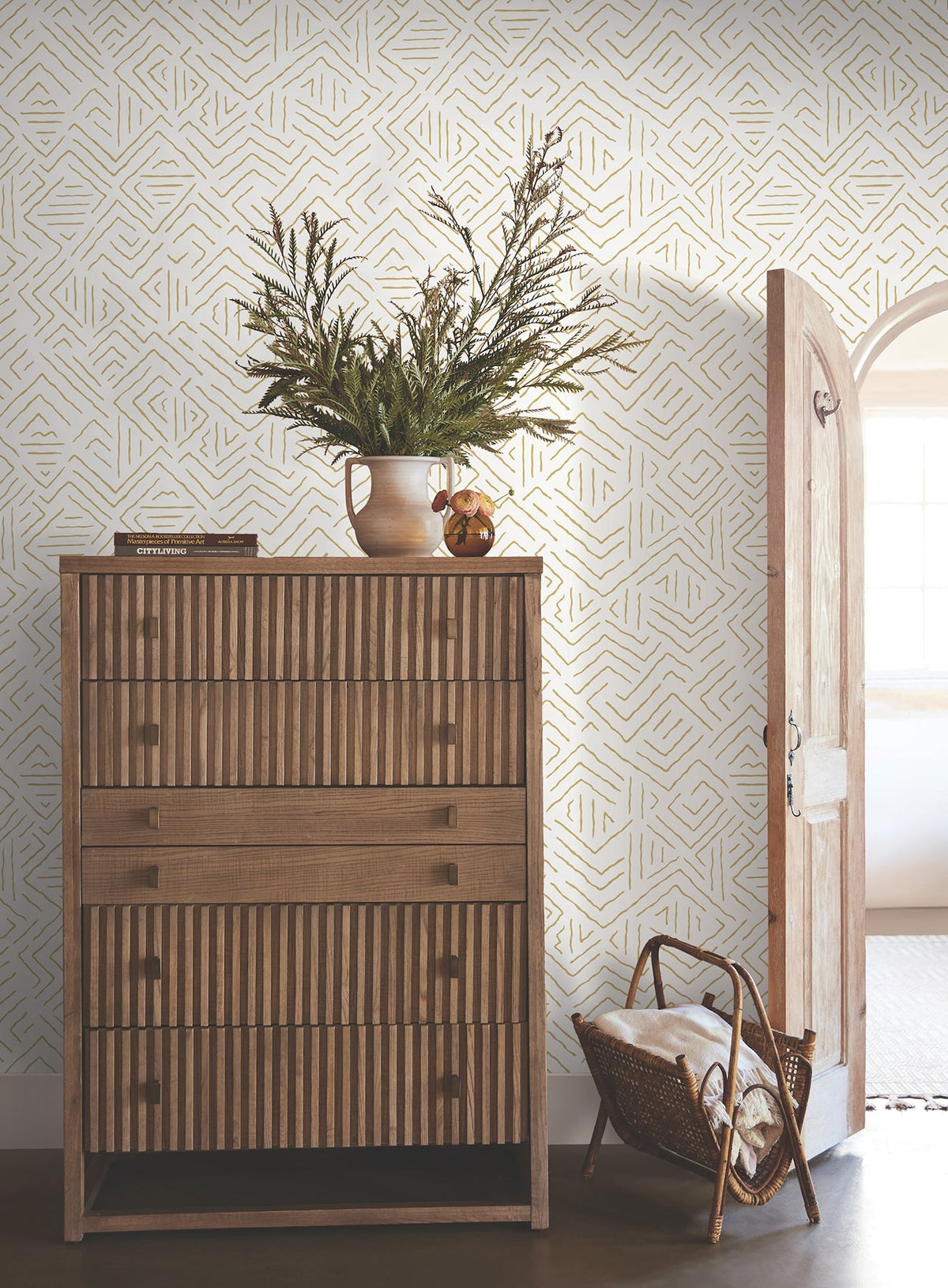 A wooden dresser with vertical grooves stands against a wall adorned with Sirih Wallpaper by York Wallcoverings. A vase with greenery and a small object sit on top. An open arched door and a wicker basket with blankets are nearby.