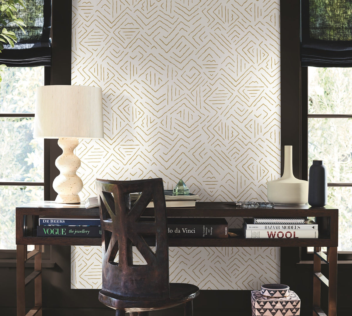 A wooden desk with books, a table lamp, a vase, and a chair sits in front of a window adorned with York Wallcoverings’ Sirih Wallpaper, featuring eye-catching geometric patterns and dark trim. Soft light filters in from outside.