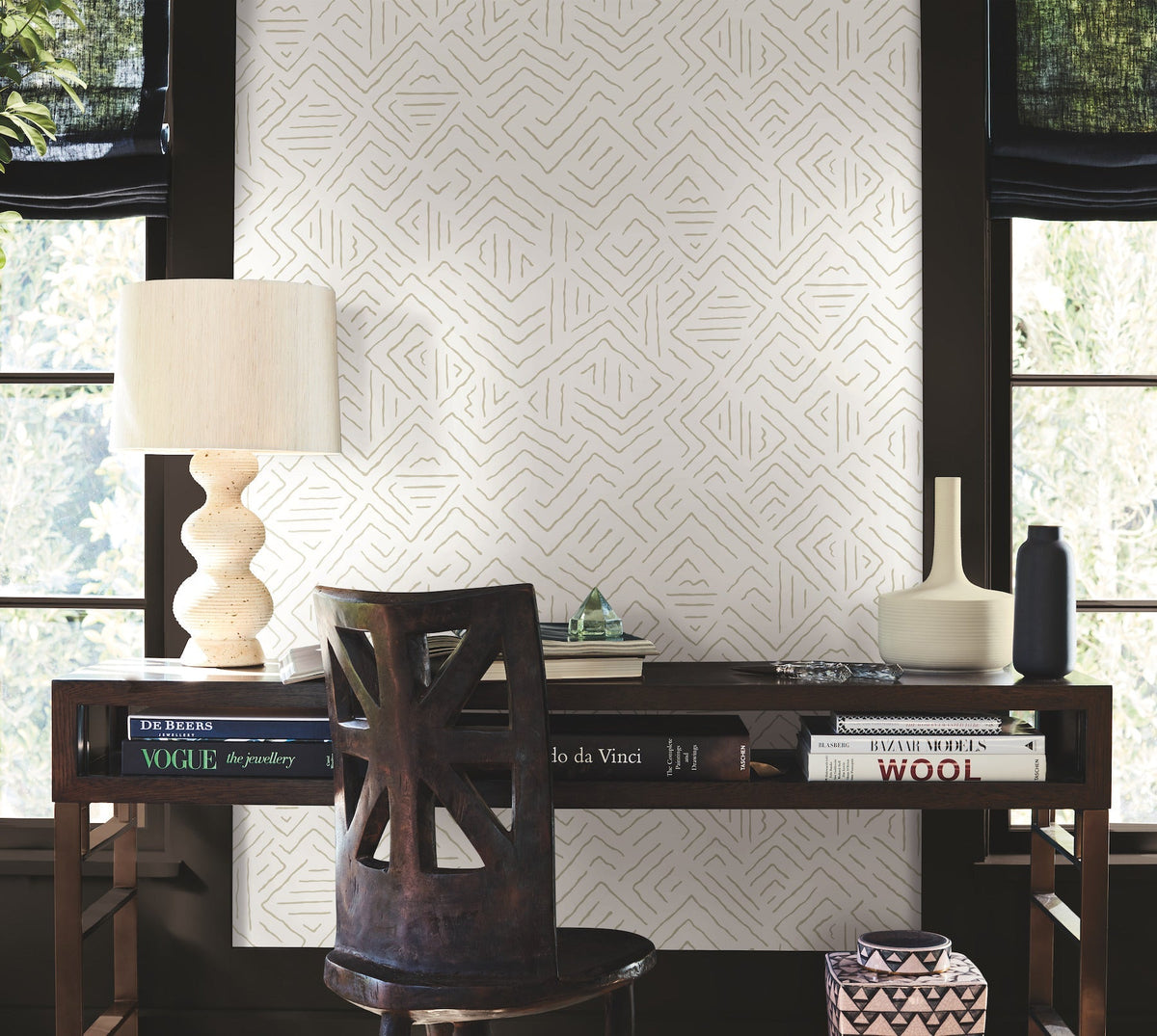 A desk with books, a lamp, decorative vases, and a chair sits by two windows. The wall is adorned with Sirih Wallpaper by York Wallcoverings featuring a geometric pattern. Natural light filters through black shades.