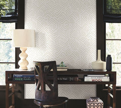 A desk with books, a lamp, decorative vases, and a chair sits by two windows. The wall is adorned with Sirih Wallpaper by York Wallcoverings featuring a geometric pattern. Natural light filters through black shades.