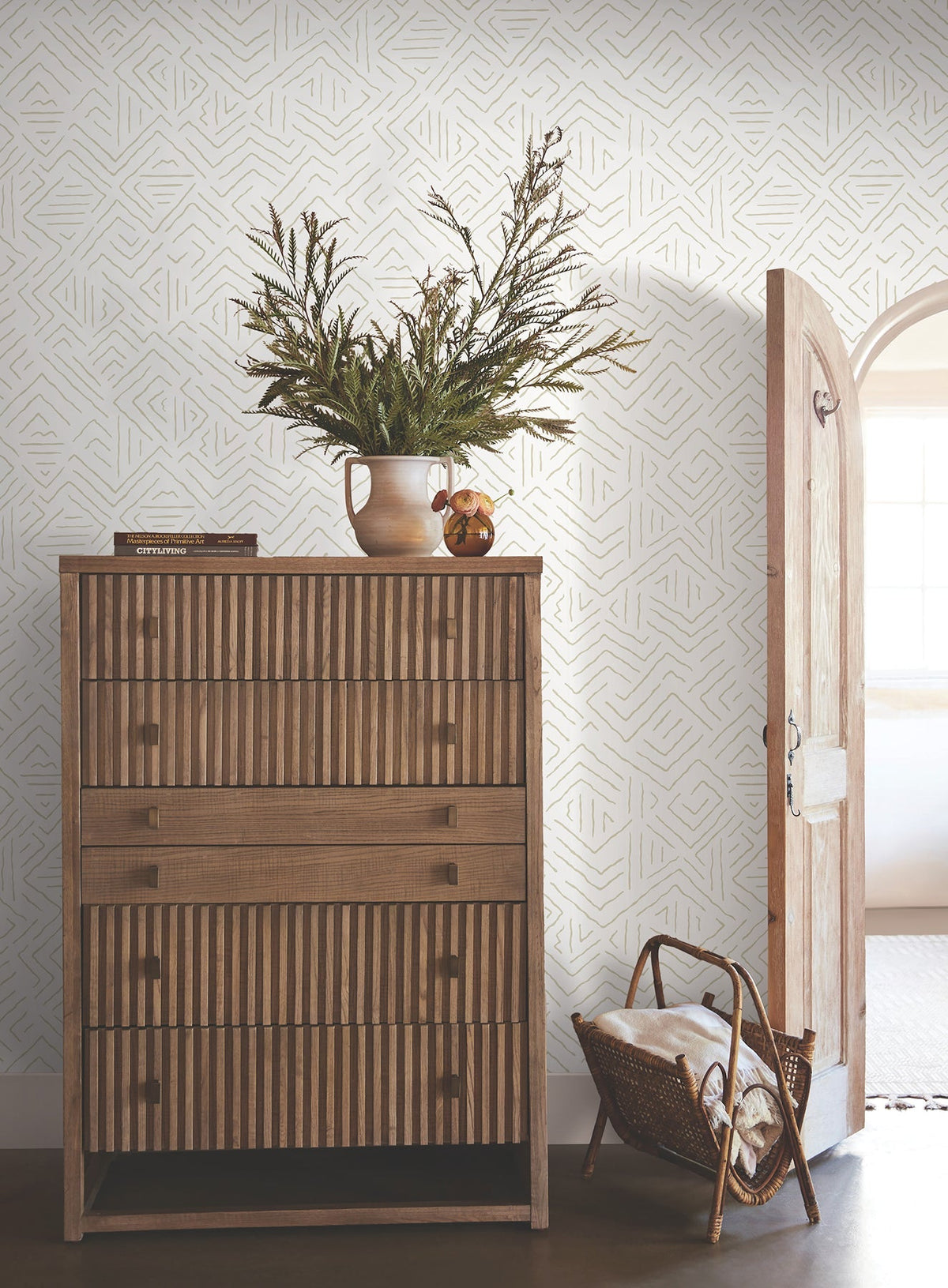 A wooden dresser with striped drawers displays a vase of greenery, books, and decorative accents. Next to it, an open arched door lets in natural light, while a woven basket filled with blankets rests on the floor. The backdrop features the Sirih Wallpaper by York Wallcoverings.