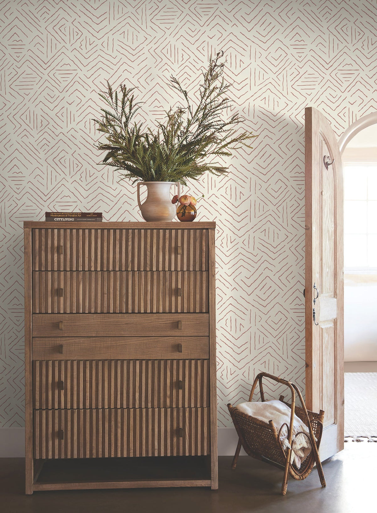 A wooden chest of drawers with striped patterns stands against a wall covered in Sirih Wallpaper by York Wallcoverings. A vase with greenery, books, and a small jar sit on top. An open arched door and a basket with towels are nearby.