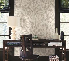 A modern desk with books, a lamp, and vases sits in front of a wall adorned with Sirih Wallpaper by York Wallcoverings, flanked by two large windows with black blinds that let in natural light.