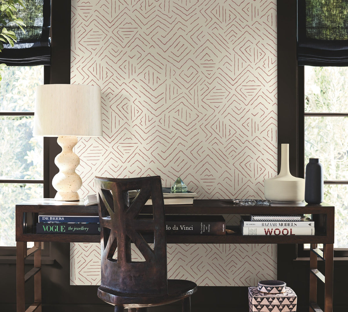 A wooden desk with books, a lamp, a vase, and a chair sits against a wall adorned with York Wallcoverings’ Sirih Wallpaper—a geometric patterned backdrop—flanked by two windows with black blinds.