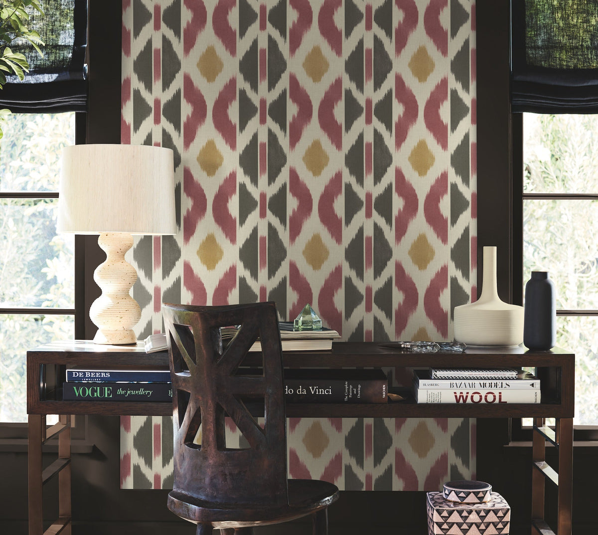 A desk adorned with books, a sculptural lamp, and a vase stands against a feature wall covered in York Wallcoverings’ Patan Wallpaper, showcasing red, gold, and white patterns. A wooden chair is positioned in front of the desk, while windows framed by dark curtains flank the wallpapered backdrop.