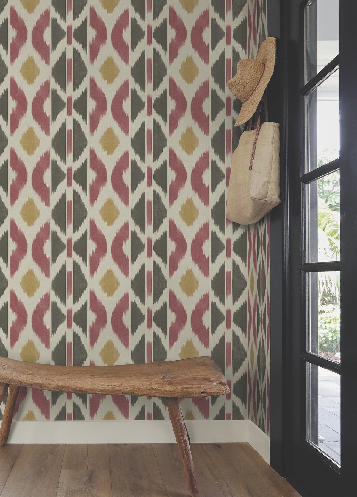 A rustic wooden bench sits by a wall adorned with the Patan Wallpaper from York Wallcoverings, featuring a striking geometric pattern. A straw hat and woven bag hang on hooks above the bench, while a glass door to the right allows sunlight to stream in from outside.