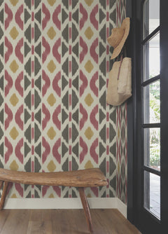 A rustic bench sits against a wall adorned with Patan Wallpaper by York Wallcoverings, featuring bold, geometric patterns in earthy tones. A straw hat and woven bag hang on a hook beside a black-framed glass door.