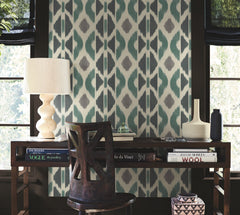A wooden desk with books, a lamp, a chair, and decorative objects sits in front of a window, set against the bold blue and gray Patan Wallpaper by York Wallcoverings.