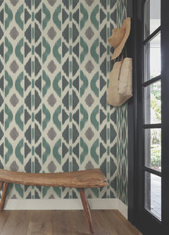 A rustic wooden bench sits against a geometric-patterned green and gray wall covered in Patan Wallpaper by York Wallcoverings. A straw hat and woven bag hang on hooks beside a glass door, with sunlight visible outside.