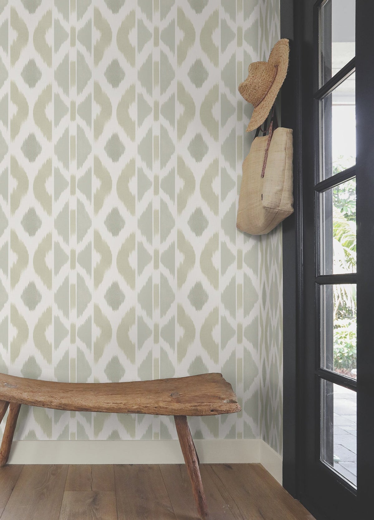 A rustic bench sits by a wall adorned with the Patan Wallpaper by York Wallcoverings, featuring a geometric pattern. Above it, a straw hat and woven bag hang beside a black-framed glass door, while wooden floorboards extend below.