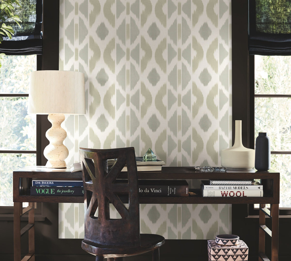 A wooden desk with books, a sculptural lamp, vases, and decor sits against a windowed wall adorned with Patan Wallpaper by York Wallcoverings. A dark wooden chair is tucked in front of the desk.
