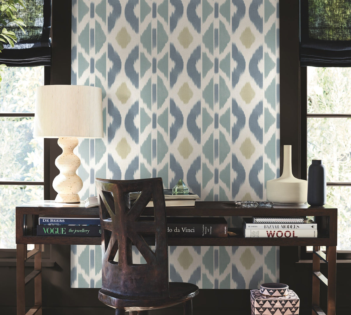 A wooden desk adorned with books, a lamp, and vases sits in front of a window featuring Patan Wallpaper by York Wallcoverings in blue and beige tones. A modern chair is pulled out, while natural light filters through black curtains.
