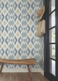 A rustic wooden bench sits in a corner adorned with Patan Wallpaper by York Wallcoverings, featuring striking geometric patterns. A straw hat and woven bag hang on a hook by the glass door, as sunlight gently filters in from outside.