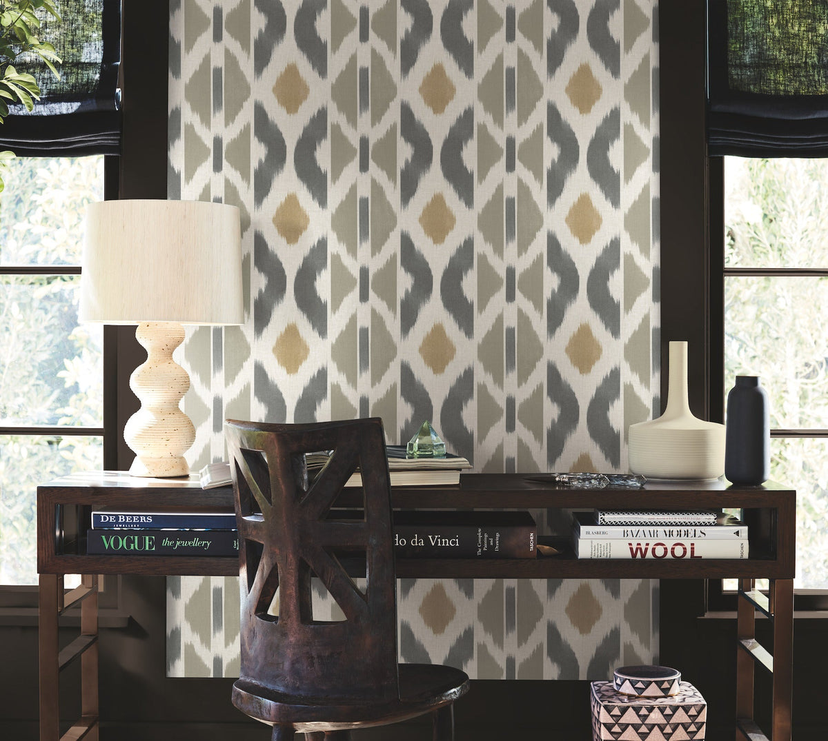 A wooden desk adorned with books, a table lamp, and vases sits in front of a window. The wall behind is decorated with Patan Wallpaper by York Wallcoverings, showcasing a gray and gold geometric pattern. Light filters in through the partially covered windows.