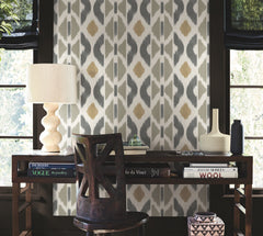A wooden desk adorned with books, a table lamp, and vases sits in front of a window. The wall behind is decorated with Patan Wallpaper by York Wallcoverings, showcasing a gray and gold geometric pattern. Light filters in through the partially covered windows.