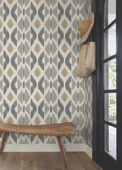 A rustic bench sits by a wall adorned with Patan Wallpaper from York Wallcoverings, featuring gentle neutral patterns. A woven hat and bag hang on hooks, while sunlight streams through a glass door, highlighting the cozy, natural decor.