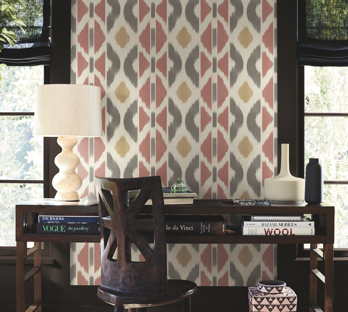 A wooden desk with books, a lamp, a chair, and decorative vases sits against a wall adorned with the Patan Wallpaper by York Wallcoverings, featuring pink, yellow, and gray geometric patterns. Sunlight filters in through windows with dark shades on both sides.