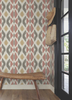A rustic wooden bench sits by a patterned wall adorned with Patan Wallpaper from York Wallcoverings, featuring pink, yellow, and gray shapes. A straw hat and woven bag hang on hooks near a black-framed glass door, where sunlight streams onto the wooden floor.