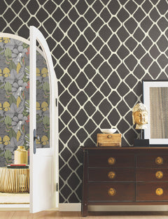 A dark dresser with a Buddha head, framed art, and an arched door opens to a room adorned with York Wallcoverings’ Tonga Wallpaper, featuring a bold black and white geometric pattern. The adjacent room features floral wallpaper.
