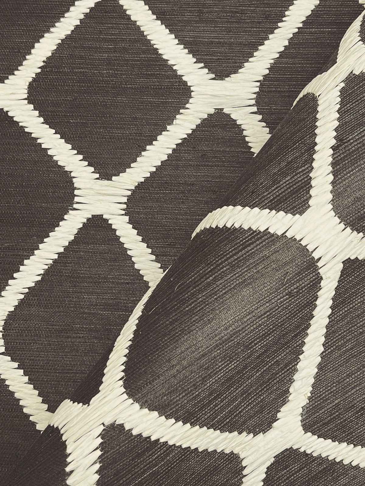 A close-up of York Wallcoverings’ Tonga Wallpaper, showcasing its dark woven texture with a white geometric diamond pattern, with one corner lifted to highlight the design and material.