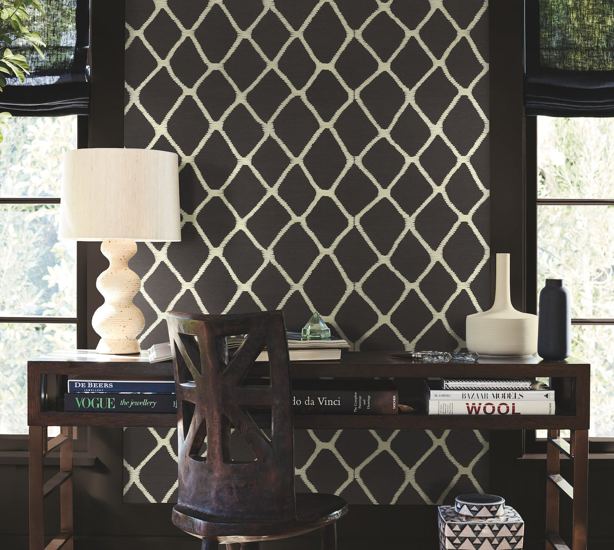 A dark wooden desk with books, a white lamp, and modern vases sits in front of a Tonga Wallpaper accent wall by York Wallcoverings, patterned in black and positioned between two windows with dark shades. A wooden chair with a cut-out back is placed in front of the desk.