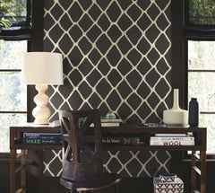 A desk with books, a lamp, vases, and a chair is arranged against an accent wall featuring Tonga Wallpaper by York Wallcoverings, set between two windows with black shades. Light filters in, illuminating the workspace.