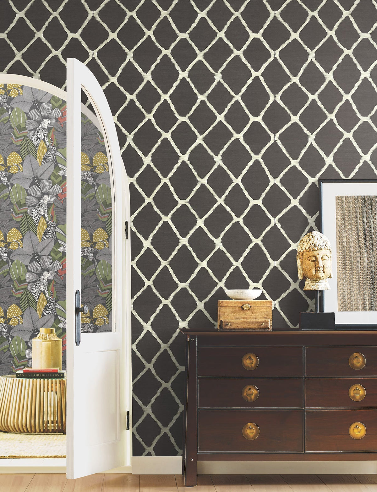 A room with dark Tonga Wallpaper by York Wallcoverings, a wooden dresser topped with a Buddha head sculpture, framed art, and an open door revealing floral wallpaper and a round gold table in the adjoining space.