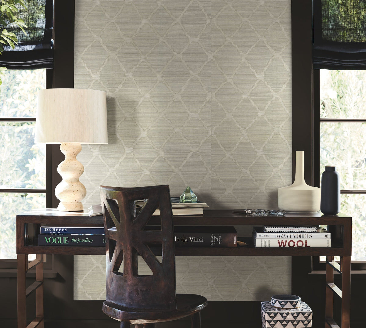 A wooden desk with books, a sculptural lamp, vases, and a chair sits in front of windows with dark trim and softly lit walls adorned in the Tonga Wallpaper by York Wallcoverings.