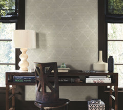 A wooden desk with books, a sculptural lamp, vases, and a chair sits in front of windows with dark trim and softly lit walls adorned in the Tonga Wallpaper by York Wallcoverings.