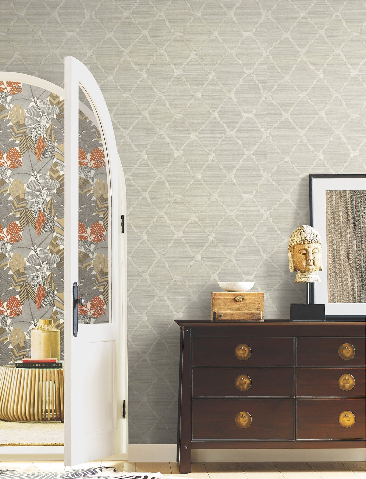 A room featuring Tonga Wallpaper by York Wallcoverings, a dark wood dresser topped with a Buddha head statue, framed art on the wall, and an open door revealing a floral-patterned interior.