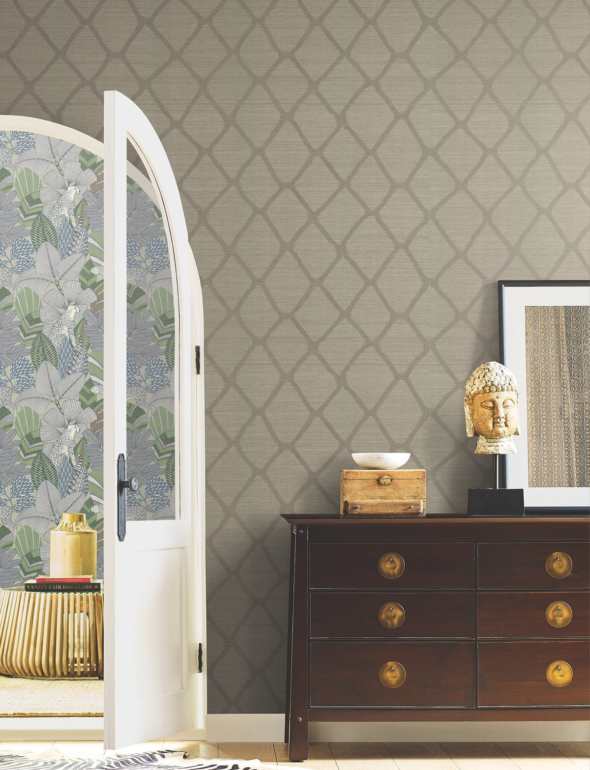 A room featuring York Wallcoverings’ Tonga Wallpaper, a dark wooden dresser, a gold Buddha head statue, framed art, and an open door revealing a floral accent wall and a round table.