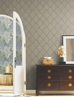 A room featuring York Wallcoverings’ Tonga Wallpaper, a dark wooden dresser, a gold Buddha head statue, framed art, and an open door revealing a floral accent wall and a round table.