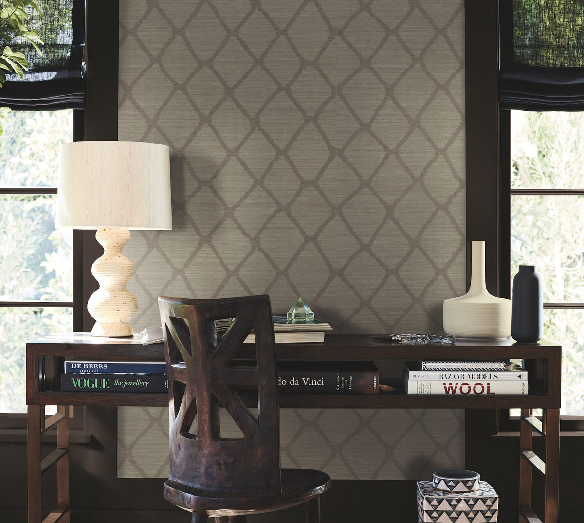 A wooden desk with books, a cream lamp, and decorative vases sits in front of two windows with black shades. A carved chair is pulled up to the desk. The wall features the Tonga Wallpaper by York Wallcoverings, showcasing a geometric pattern.