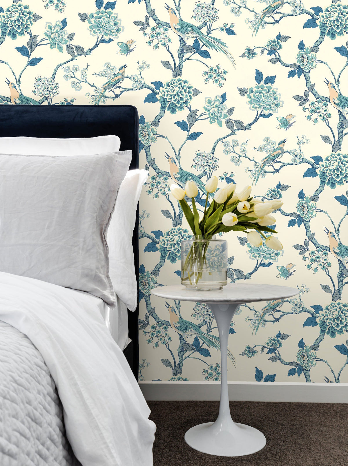 A bedside table with a glass vase of white tulips stands next to a bed with white linens, set against York Wallcoverings’ Fanciful Wallpaper featuring blue birds and flowers.