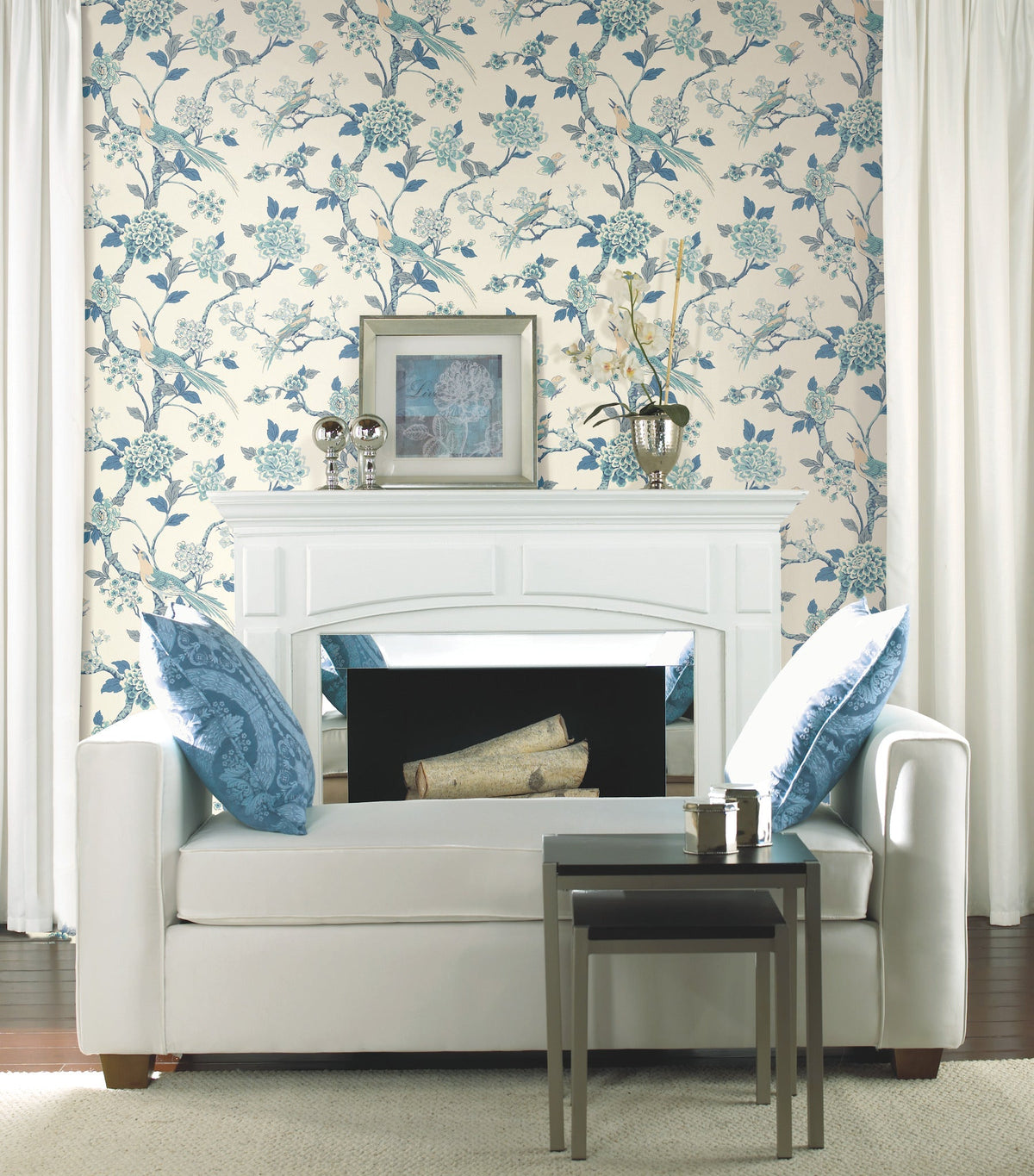 A white sofa with blue-patterned pillows sits in front of a white fireplace. “Fanciful Wallpaper” by York Wallcoverings in floral blue and cream hues, along with white curtains, frames the scene. A framed picture, vases, and logs decorate the mantel and fireplace.