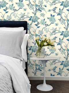 A bed with white linens sits next to a small round white table displaying a glass vase of white tulips, set against York Wallcoverings’ Fanciful Wallpaper featuring blue flowers and birds.