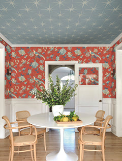 A dining area features a round white table surrounded by four wooden chairs, topped with a vase of leafy branches and lemons. The walls are adorned with York Wallcoverings’ Fanciful Wallpaper in a vibrant red floral pattern, while the blue patterned ceiling adds contrast. White doors and a wood floor complete the room.