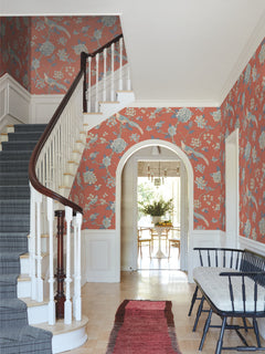 A hallway features a curved staircase, a black bench, and a red rug, with the walls adorned in York Wallcoverings’ Fanciful Wallpaper in coral floral. An arched doorway opens to a dining area furnished with a table, chairs, and a vase of greenery.