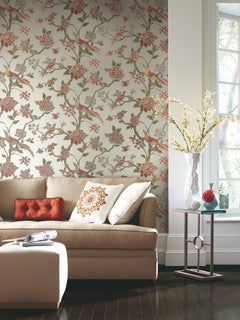 A beige sofa with decorative pillows sits in front of a wall adorned with York Wallcoverings’ Fanciful Wallpaper. A small table holds a white vase filled with yellow flowers, while sunlight streams through a nearby window.