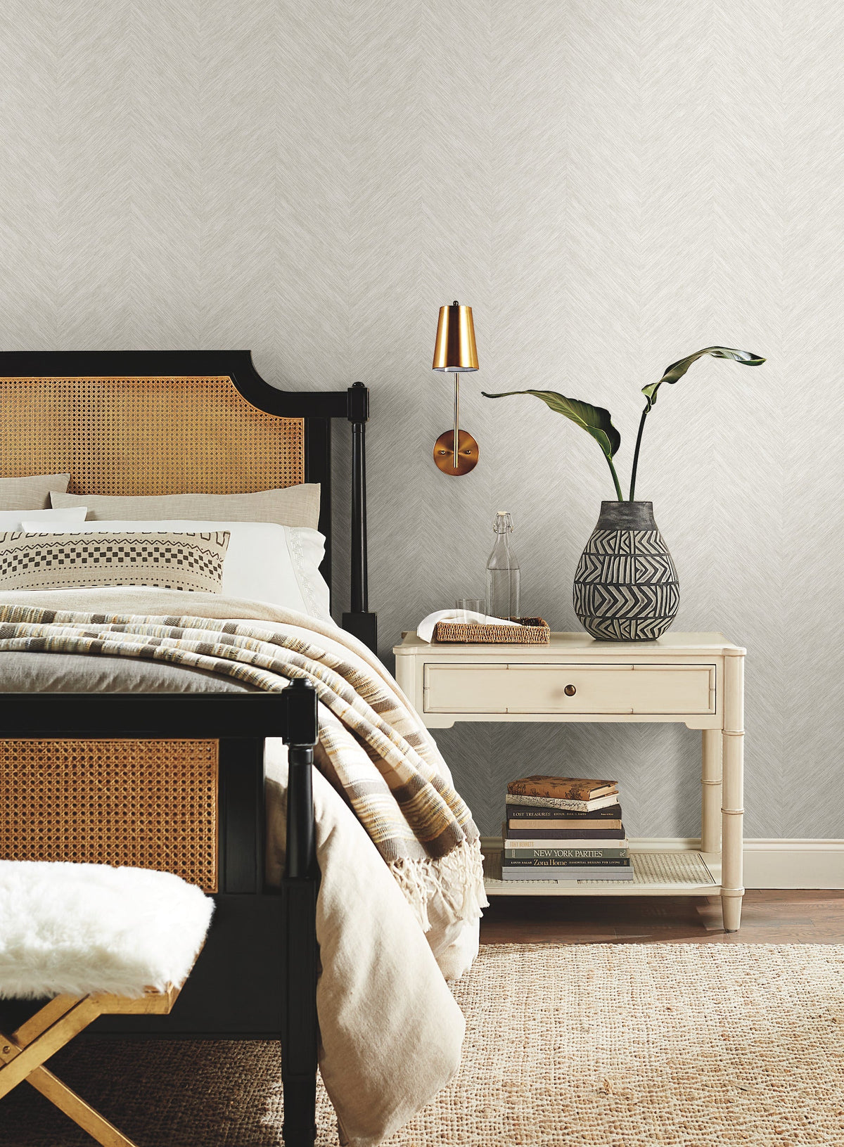A cozy bedroom featuring York Wallcoverings’ Chevron Wallpaper, complemented by a black and wicker bed with white bedding, a beige-striped throw, a white nightstand, a patterned vase filled with green leaves, stacked books, and a gold wall sconce.
