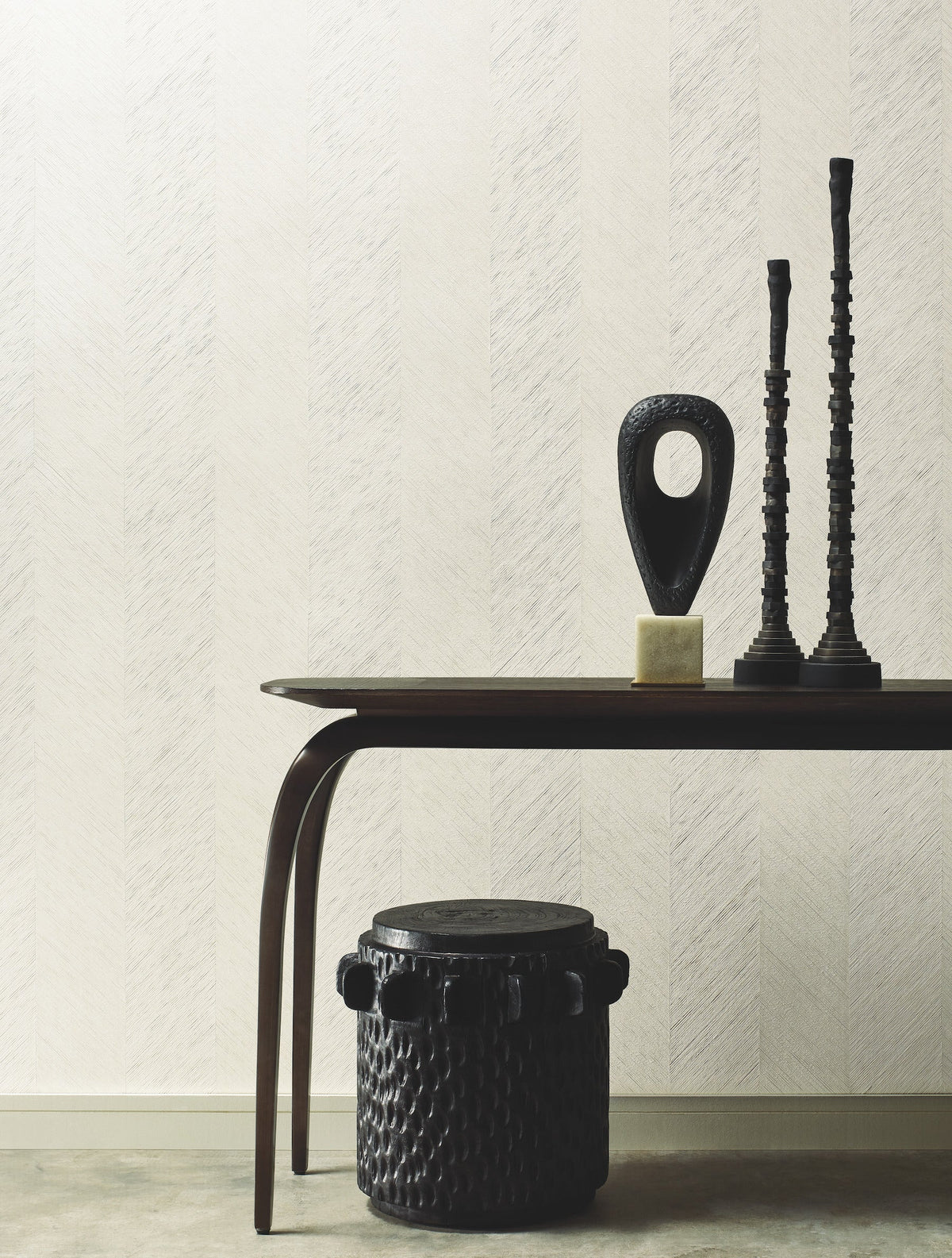 A modern wooden table adorned with abstract black sculptures and candlesticks stands against a textured cream wall covered in York Wallcoverings’ Chevron Wallpaper. Below, a round, black, textured stool or drum rests on the stone floor. The scene is minimal and stylish.