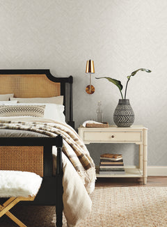 A cozy bedroom featuring York Wallcoverings’ Chevron Wallpaper, a black and cane bed, striped bedding, and a beige nightstand topped with a patterned vase of green leaves and books, all illuminated by a modern brass wall sconce above.