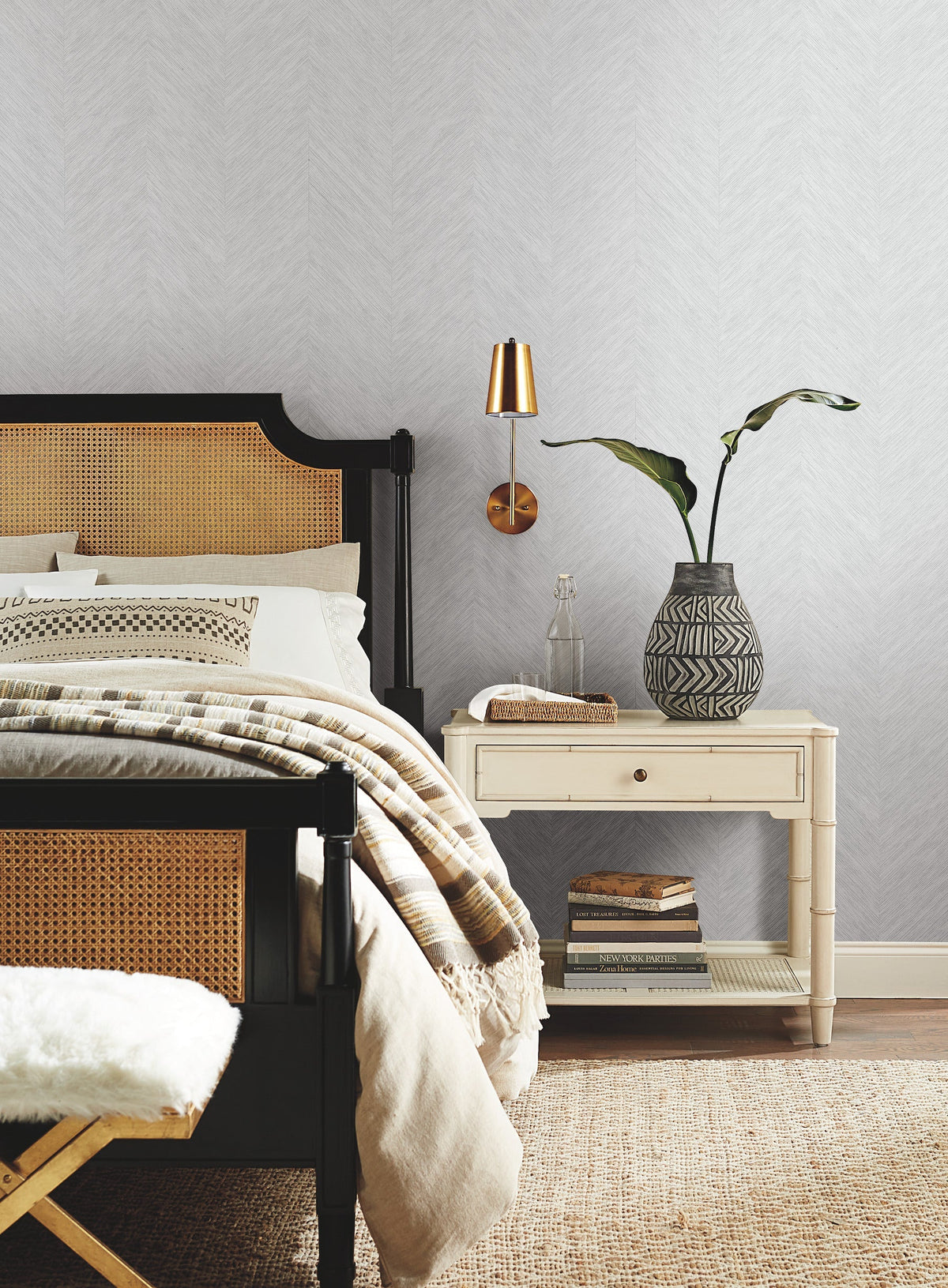 A stylish bedroom features York Wallcoverings’ Chevron Wallpaper as a backdrop for a black and cane bed with beige bedding, paired with a white nightstand holding a patterned vase with a plant, books, and accented by a gold wall sconce.